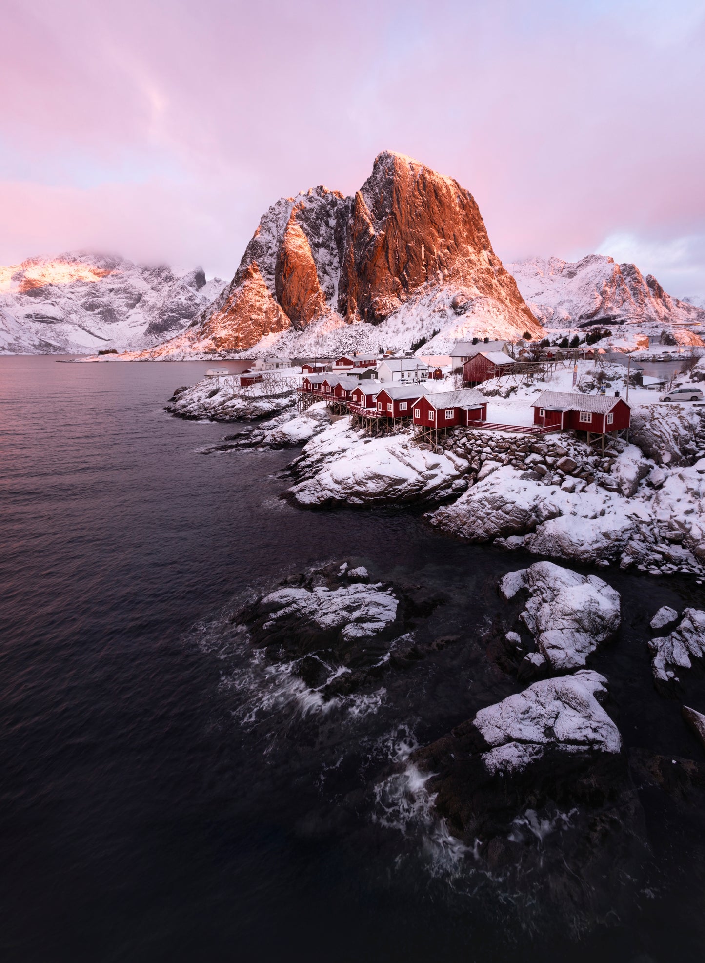 Hamnøy fishing village in Norway's Lofoten Islands at dusk, featuring traditional red rorbuer cabins reflected in calm Arctic waters with dramatic mountain peaks in the background - fine art landscape photography print